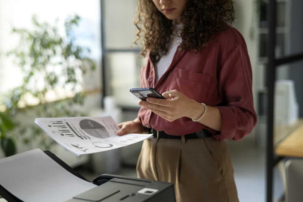 Conheça 5 novas tecnologias de impressão e adote na sua empresa 9 Mulher usando o smartphone enquanto retira um documento impresso de uma impressora