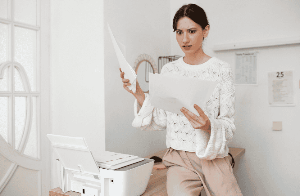 Mulher com expressão de preocupação segurando folhas de papel ao lado de uma impressora