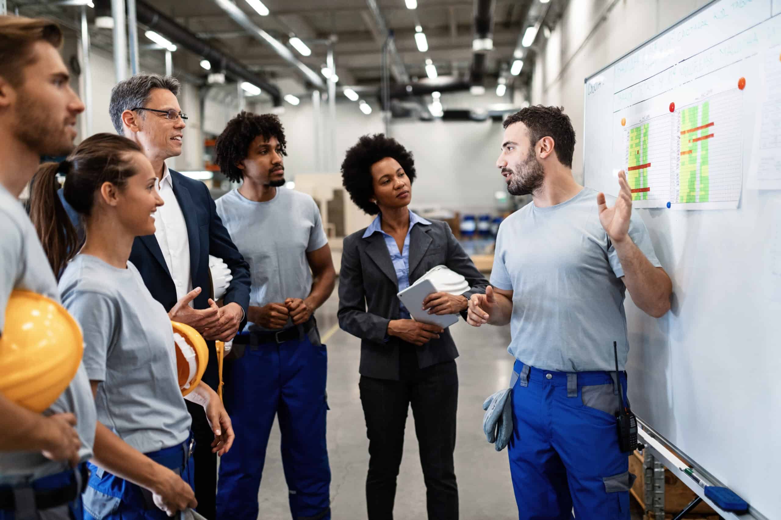 Equipe reunida diante de um quadro com gr&aacute;ficos durante apresenta&ccedil;&atilde;o de estrat&eacute;gia em uma f&aacute;brica