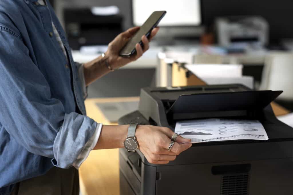 Pessoa segurando um smartphone e retirando um documento impresso de uma impressora