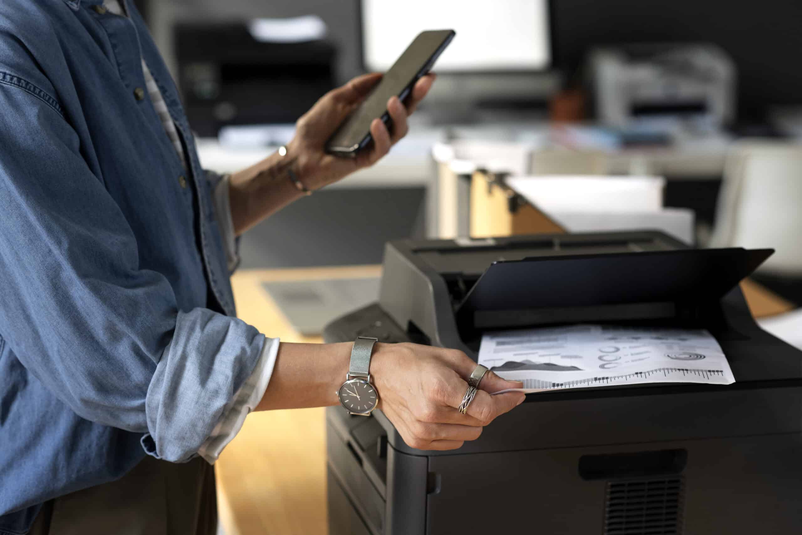 Pessoa segurando um smartphone e retirando um documento impresso de uma impressora